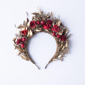 May include: A decorative headband featuring red roses and berries, accented with gold-colored leaves and foliage. The floral crown is set against a white background, showcasing its intricate design and textures. This headpiece is suitable for special occasions.