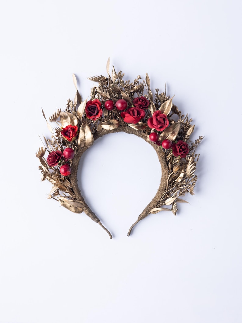 May include: A decorative headband featuring red roses and berries, accented with gold-colored leaves and foliage. The floral crown is set against a white background, showcasing its intricate design and textures. This headpiece is suitable for special occasions.