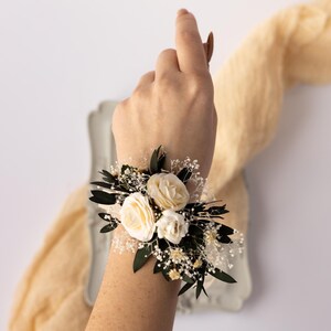 Peut inclure: Une corsage de poignet avec des fleurs blanches et de la verdure. La corsage est attachée avec un ruban blanc et comprend de petites fleurs blanches et des feuilles vertes.