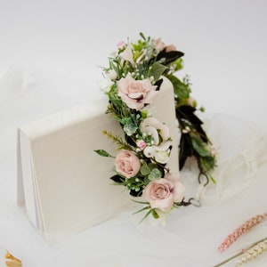 May include: A delicate floral crown with pink and white flowers and green leaves. The crown is resting on a white surface.