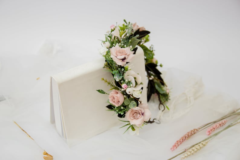 May include: A delicate floral crown with pink and white flowers and green leaves. The crown is resting on a white surface.