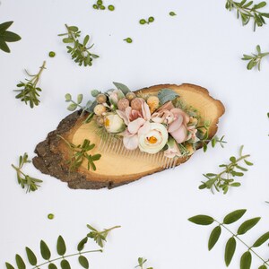 May include: A floral hair comb with pink and white flowers and greenery, displayed on a wooden slice.