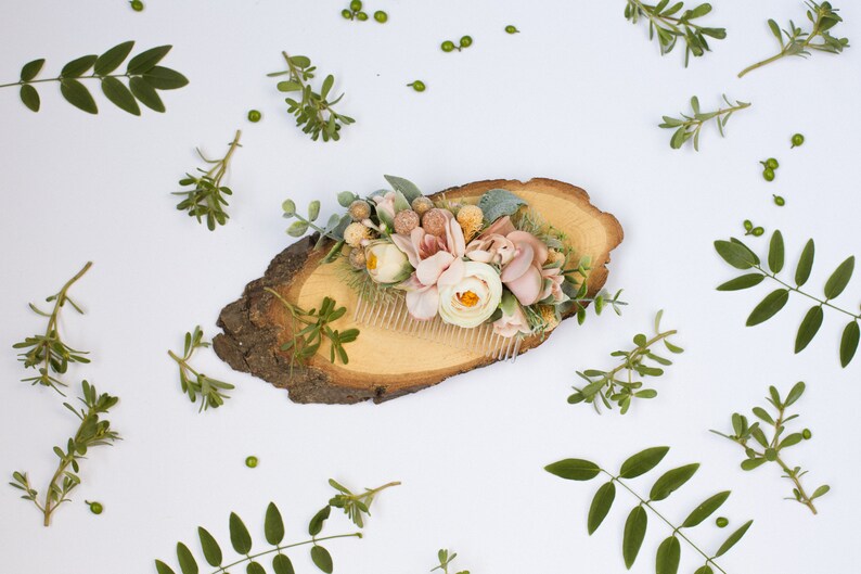 May include: A floral hair comb with pink and white flowers and greenery, displayed on a wooden slice.