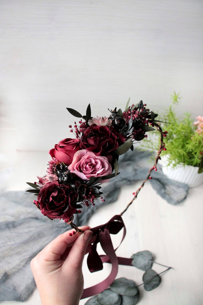 May include: A burgundy floral crown featuring pink roses and burgundy berries. The crown is crafted with a brown wire base and has a burgundy ribbon tied at the bottom.