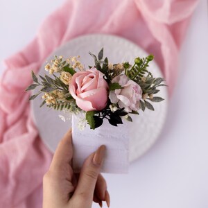 Könnte beinhalten: Ein kleiner Strauß mit rosa Rosen und Grün, der in einer weißen Keramikvase arrangiert ist. Der Strauß wird in einer Hand gehalten.