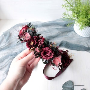 May include: A burgundy floral crown with a burgundy ribbon. The crown is made of dried flowers and greenery, including roses, eucalyptus leaves, and berries.