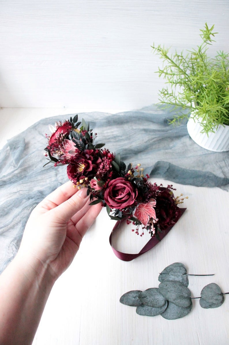 May include: A burgundy floral crown with a burgundy ribbon. The crown is made of dried flowers and greenery, including roses, eucalyptus leaves, and berries.