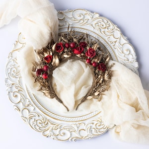 May include: A decorative headband featuring red roses and berries, accented with gold-colored leaves and stems. The floral crown is set on a brown base and displayed on a white, ornate plate with a cream-colored fabric draped around it.
