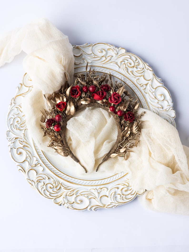 May include: A decorative headband featuring red roses and berries, accented with gold-colored leaves and stems. The floral crown is set on a brown base and displayed on a white, ornate plate with a cream-colored fabric draped around it.