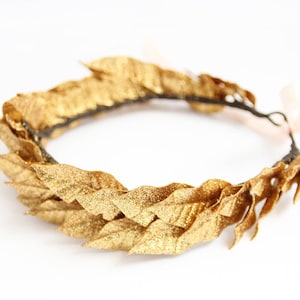 May include: A gold laurel wreath headband with glitter accents. The headband is made of a flexible wire and is decorated with gold leaves.