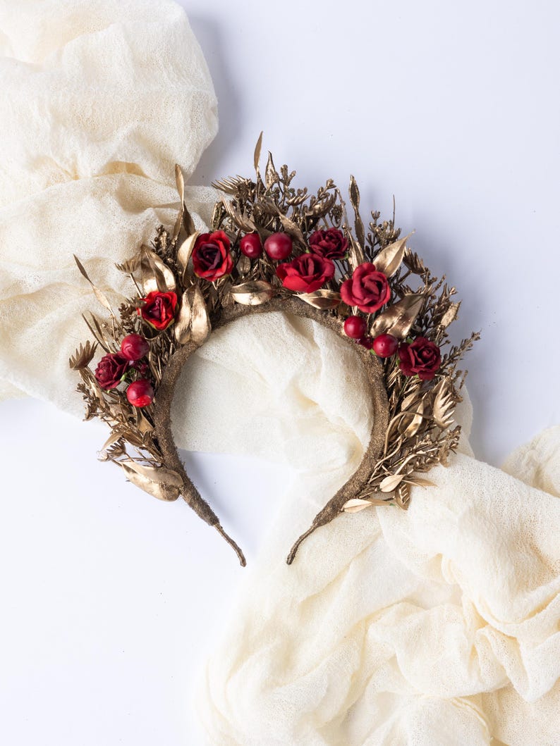 May include: A handcrafted floral crown featuring red roses and berries, accented with gold-colored leaves and foliage. The crown is set on a brown headband, perfect for special occasions or themed events.