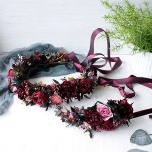 May include: Two burgundy floral crowns with dark green leaves and a pink rose. The crowns are on a white surface with green leaves scattered around them.