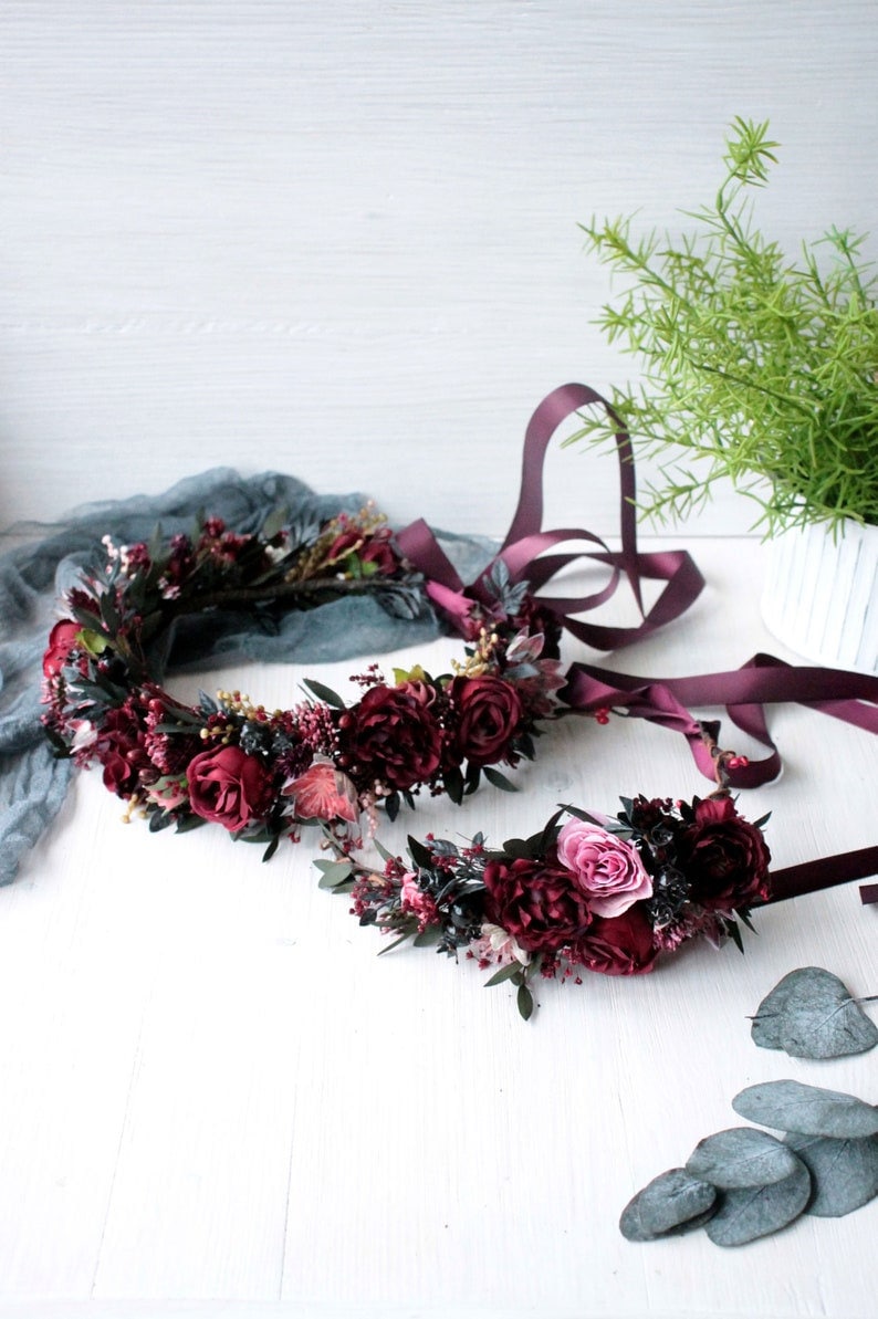 May include: Two burgundy floral crowns with dark green leaves and a pink rose. The crowns are on a white surface with green leaves scattered around them.