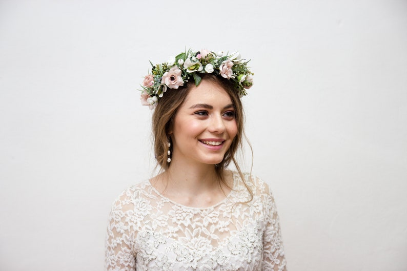 May include: A woman wearing a white lace dress with a floral crown on her head. The crown is made of pink and white flowers and greenery.