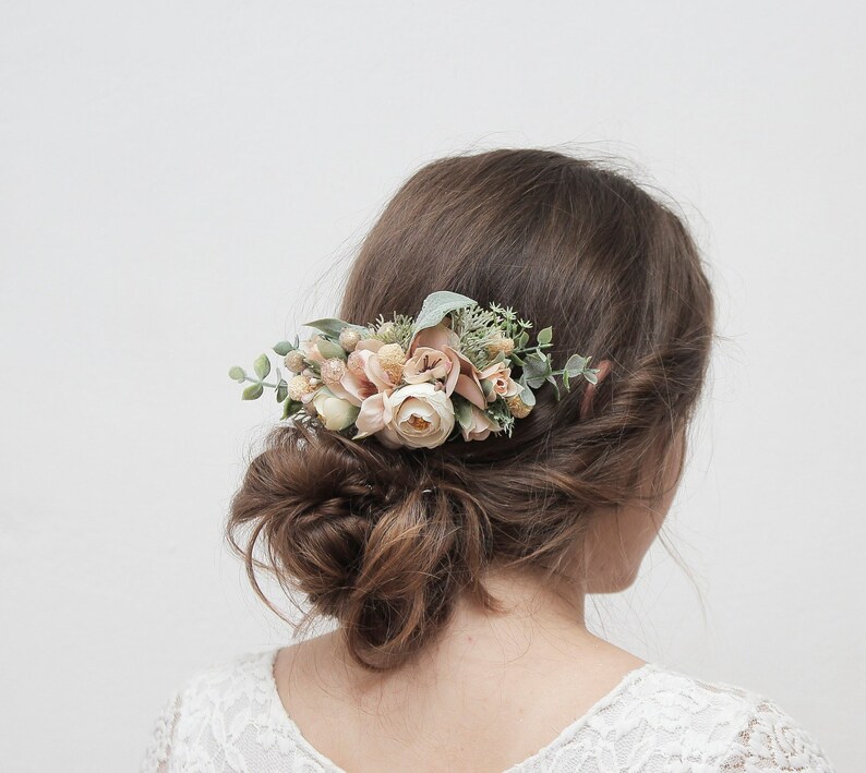May include: A floral hair comb with blush pink and white flowers and greenery. The comb is worn in a woman's hair.