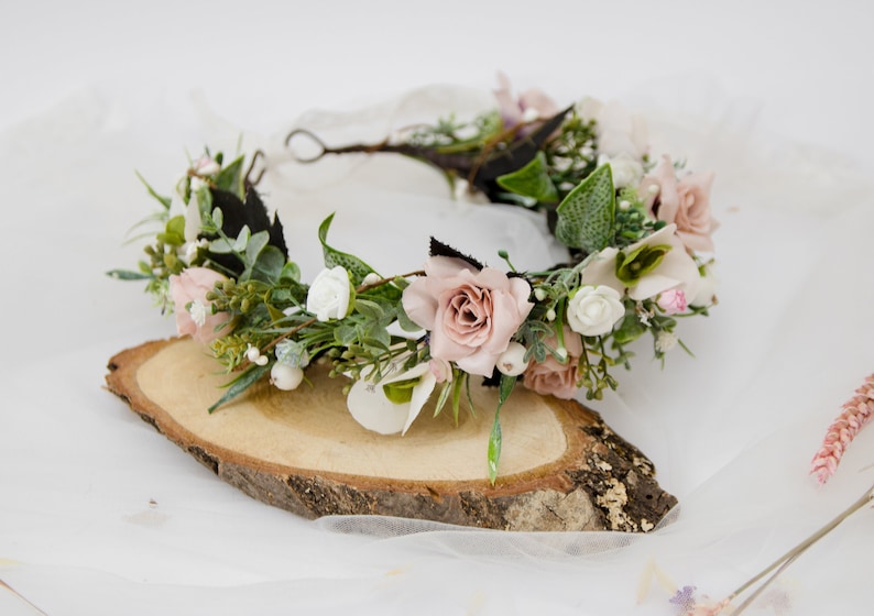 May include: A floral crown with pink and white flowers, green leaves, and a brown wire base. The crown is resting on a wooden slice.