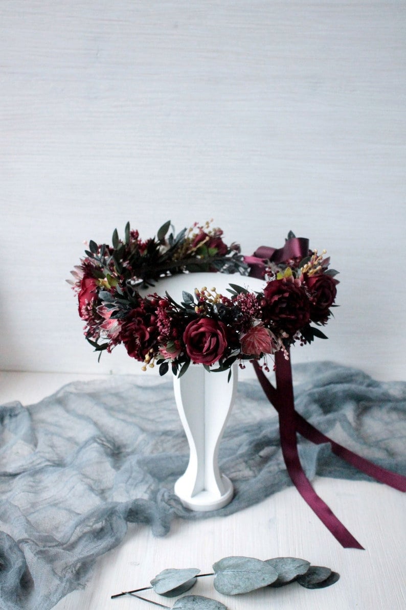 May include: Burgundy floral crown with a burgundy ribbon. The crown is made of silk flowers and greenery and is displayed on a white stand.