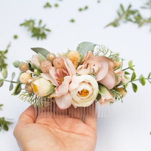 May include: A blush pink and cream floral hair comb with greenery and gold accents. The comb is held in a hand.