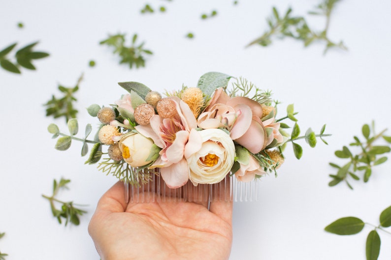 May include: A blush pink and cream floral hair comb with greenery and gold accents. The comb is held in a hand.