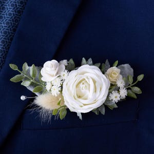 Peut inclure: Une boutonnière florale blanche avec une grande rose et de petites fleurs, de la verdure et un accent de perle, présentée sur une veste de costume bleu marine. L'arrangement est dans la poche de la veste, créant un look classique et élégant.