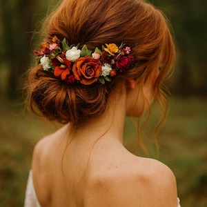 May include: A woman's auburn hair is styled in a low bun, adorned with a floral hairpiece. The hairpiece features orange, red, white, and yellow flowers. The woman is wearing a white lace dress, with the back of her shoulders visible.