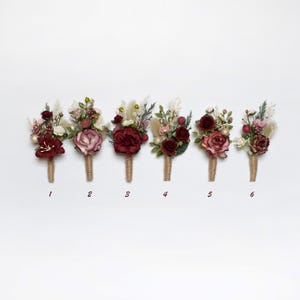 Pode incluir: Seis boutonnières florais de cor bordô e branco com hastes de juta. As boutonnières são numeradas de 1 a 6.