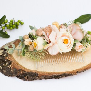 May include: A delicate floral hair comb with blush pink and white flowers, greenery, and gold accents. The comb is displayed on a wooden slice.