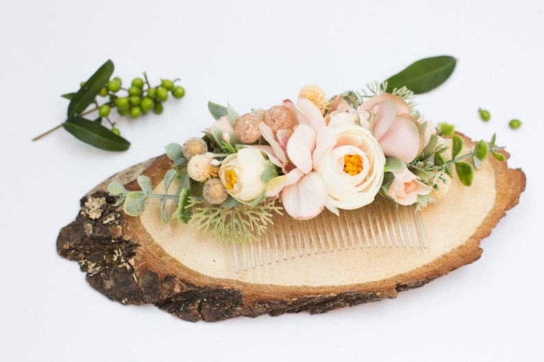 May include: A delicate floral hair comb with blush pink and white flowers, greenery, and gold accents. The comb is displayed on a wooden slice.