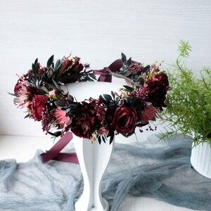 May include: A burgundy floral crown with dark green leaves and a burgundy ribbon. The crown is displayed on a white pedestal.