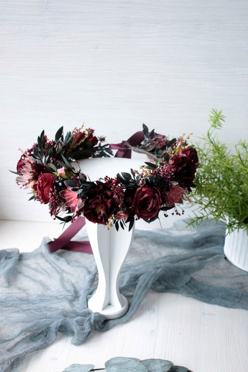 May include: A burgundy floral crown with dark green leaves and a burgundy ribbon. The crown is displayed on a white pedestal.