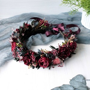 May include: A burgundy floral crown with a burgundy satin ribbon. The crown is made with burgundy roses, burgundy berries, and green leaves.