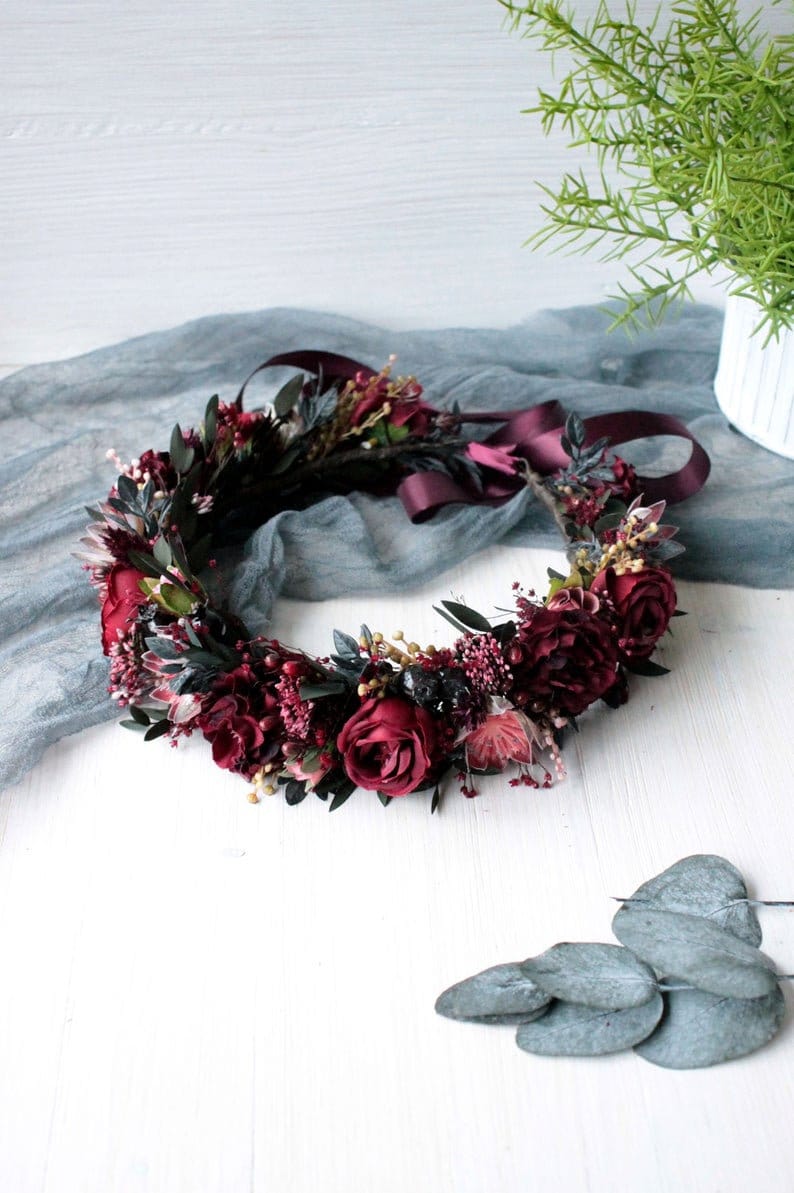 May include: A burgundy floral crown with a burgundy satin ribbon. The crown is made with burgundy roses, burgundy berries, and green leaves.