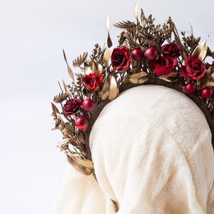 May include: A decorative floral crown featuring red roses and berries, accented with gold-colored leaves and foliage. The crown is set against a white background, showcasing its intricate design and details. This headpiece is suitable for special occasions.