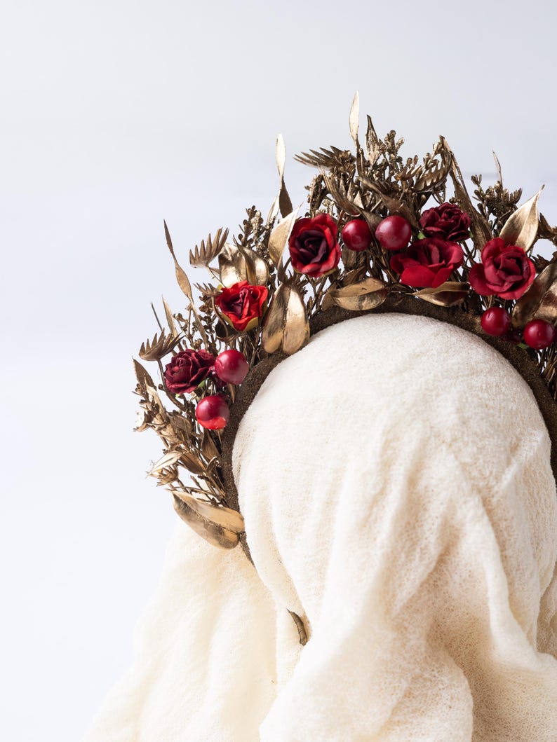 May include: A decorative floral crown featuring red roses and berries, accented with gold-colored leaves and foliage. The crown is set against a white background, showcasing its intricate design and details. This headpiece is suitable for special occasions.