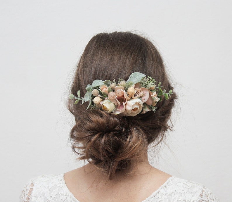 May include: A floral hair comb with blush pink and white flowers and greenery. The comb is worn in a woman's hair.