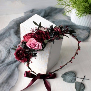 May include: A burgundy floral crown with a burgundy satin ribbon. The crown is made of twigs and has small red berries. The crown is lying on a white surface with a white box and green leaves.