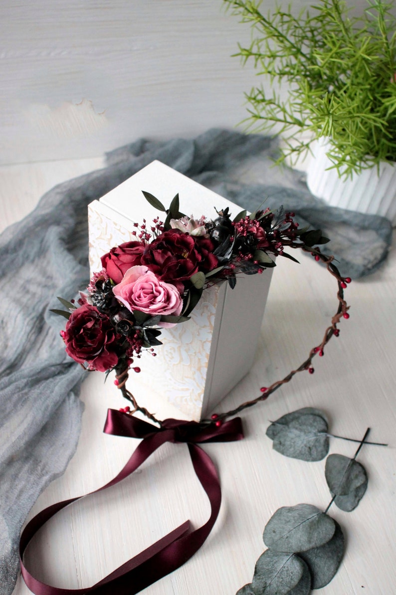 May include: A burgundy floral crown with a burgundy satin ribbon. The crown is made of twigs and has small red berries. The crown is lying on a white surface with a white box and green leaves.