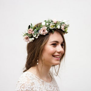 May include: A woman wearing a white lace dress with a floral crown made of pink and white flowers and greenery. She is smiling and looking to the right.