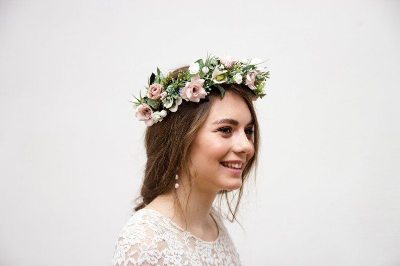 May include: A woman wearing a white lace dress with a floral crown made of pink and white flowers and greenery. She is smiling and looking to the right.