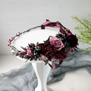 May include: A burgundy and pink floral crown with a burgundy ribbon. The crown is made of faux flowers and greenery and is sitting on a white pedestal.