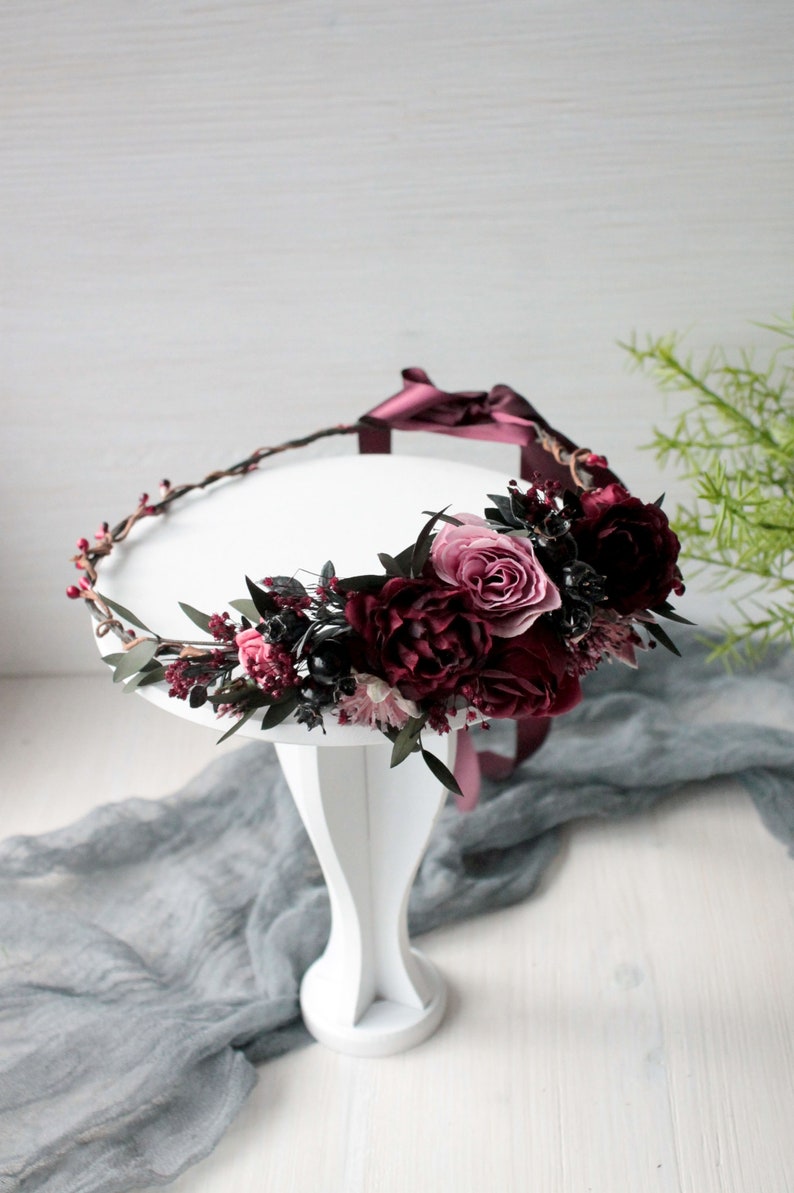 May include: A burgundy and pink floral crown with a burgundy ribbon. The crown is made of faux flowers and greenery and is sitting on a white pedestal.