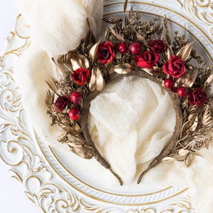 May include: A decorative headpiece featuring a brown base adorned with gold-colored leaves and red roses. The headpiece is embellished with red berries and rests on a white, ornate plate with gold trim. A cream-colored fabric is draped around the headpiece.
