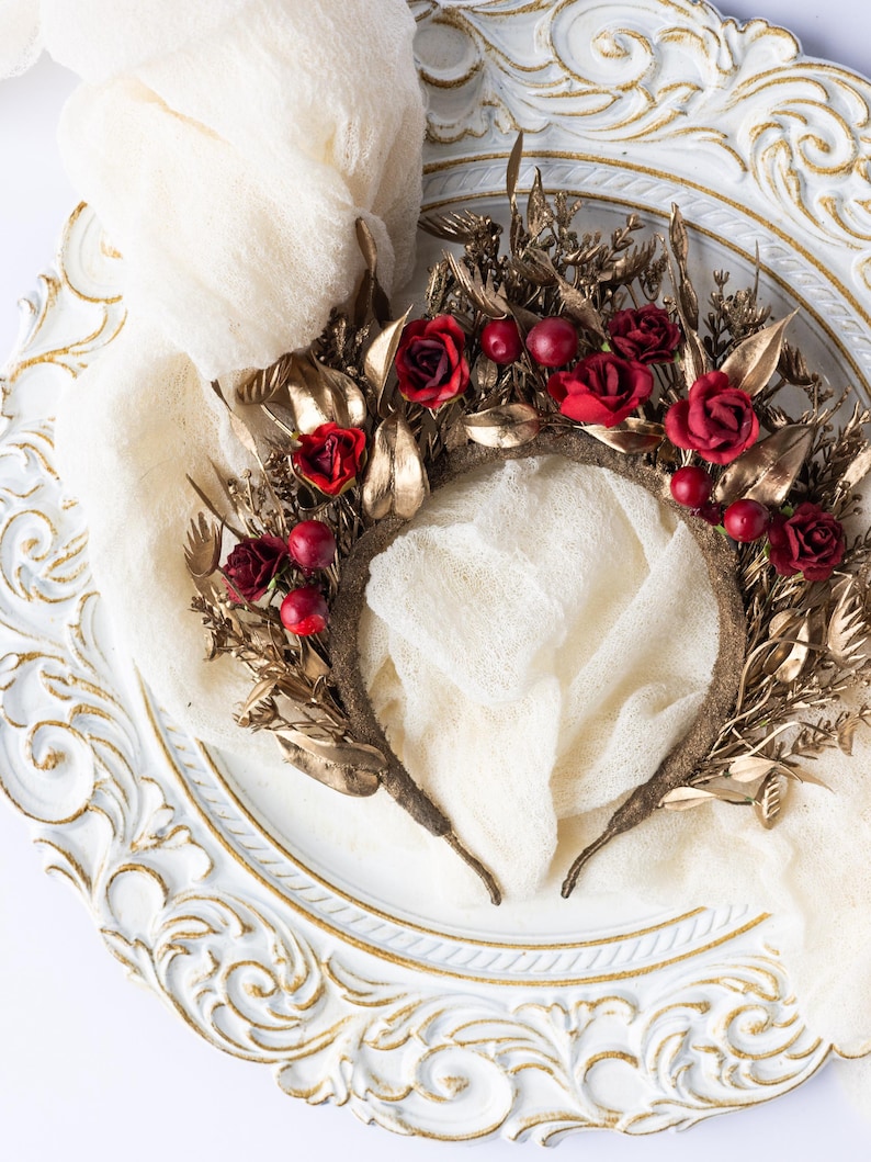 May include: A decorative headpiece featuring a brown base adorned with gold-colored leaves and red roses. The headpiece is embellished with red berries and rests on a white, ornate plate with gold trim. A cream-colored fabric is draped around the headpiece.