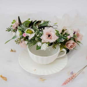 May include: A delicate floral crown with pink and white flowers, green leaves, and a black headband, resting on a white teacup and saucer.