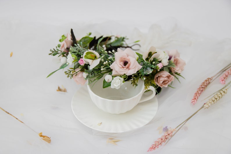 May include: A delicate floral crown with pink and white flowers, green leaves, and a black headband, resting on a white teacup and saucer.