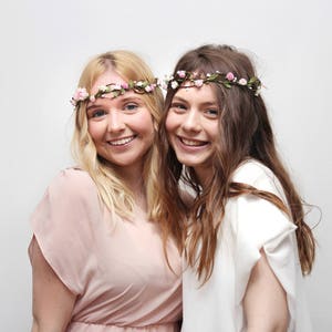 May include: Two women wearing floral crowns, one with a pink dress and the other with a white dress. They are smiling and looking at the camera.
