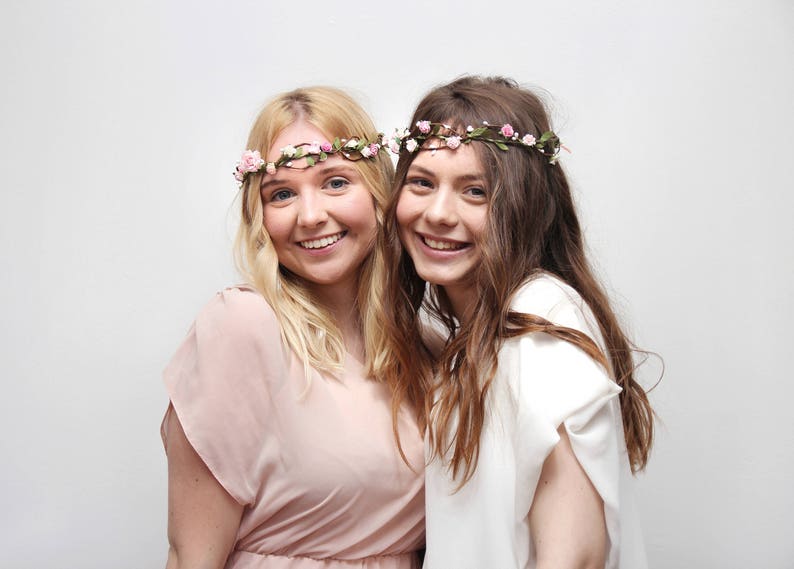 May include: Two women wearing floral crowns, one with a pink dress and the other with a white dress. They are smiling and looking at the camera.