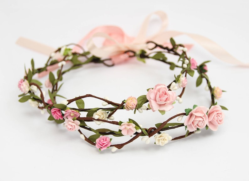 May include: A double-layered floral crown with pink and white flowers on brown branches. The crown is made of artificial flowers and is tied with a pink ribbon.