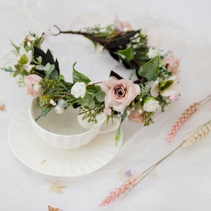 May include: A delicate floral crown with pink and white flowers, green leaves, and a brown wire base. The crown is lying on a white surface with a white teacup and saucer.