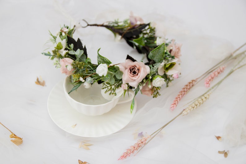 May include: A delicate floral crown with pink and white flowers, green leaves, and a brown wire base. The crown is lying on a white surface with a white teacup and saucer.